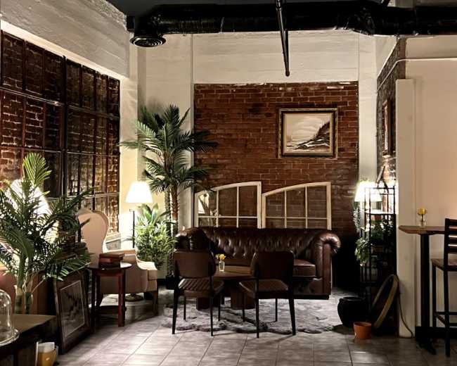 The image features a cozy seating area in a rustic cafe, with a brown leather sofa, wooden chairs, and decorative plants amidst exposed brick walls.