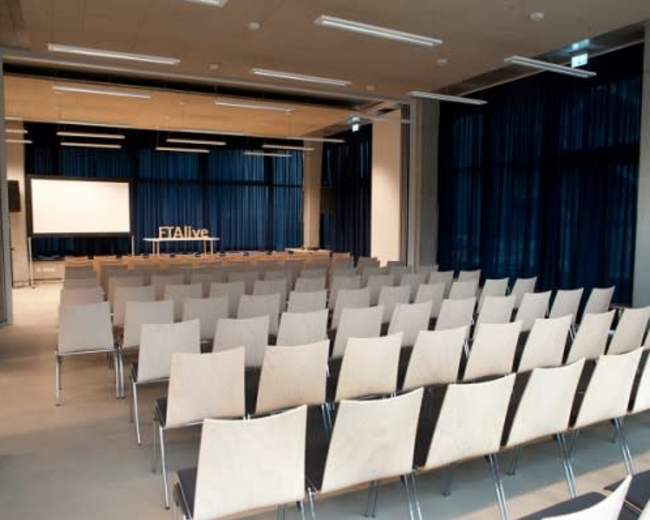 A spacious conference room with rows of empty wooden chairs facing a large screen and a backdrop with the text "#Alive."