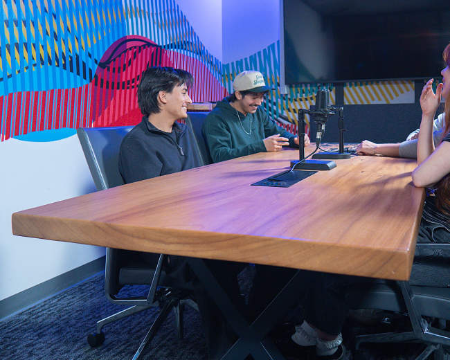 A group of four individuals is seated around a wooden conference table in a brightly colored meeting room, with microphones positioned in front of them.