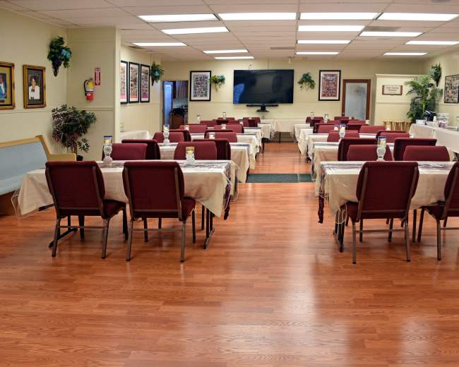 A large dining area features rows of tables covered with light-colored tablecloths and chairs arranged around them, while walls are adorned with framed photographs.