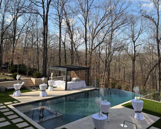 The image shows a spacious outdoor area featuring a rectangular swimming pool, surrounded by several white-topped tables and wooded scenery in the background.