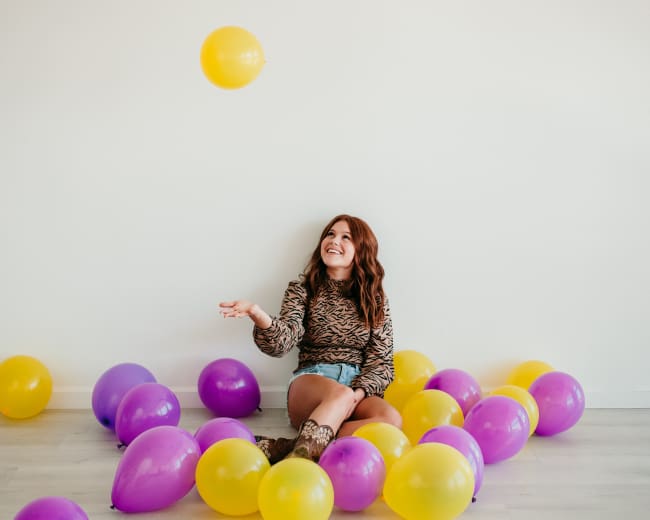 A woman sits on the floor surrounded by purple and yellow balloons, playfully tossing a yellow balloon into the air.