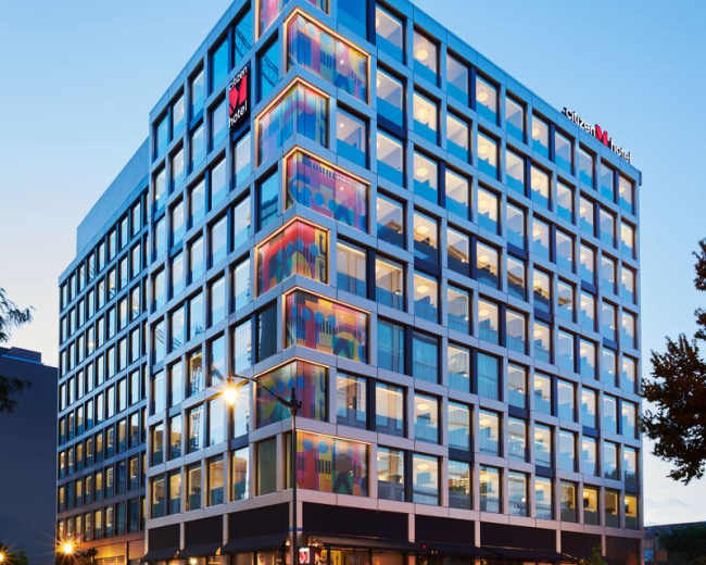 A modern multi-story hotel building with a glass facade and colorful accents at the corners, situated on a city street.