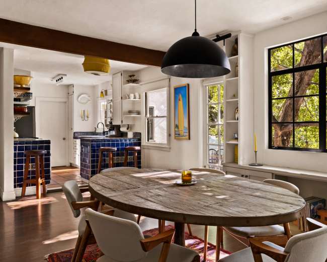 The image shows a bright kitchen and dining area featuring a round wooden table, modern chairs, and large windows that provide natural light and views of outdoor greenery.
