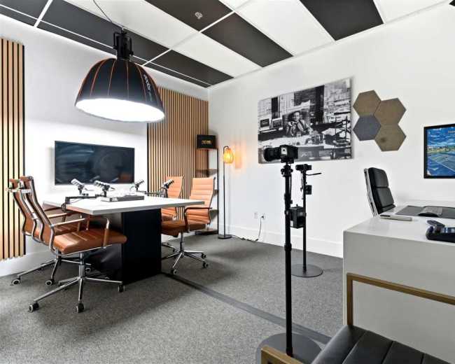 The image shows a modern office space featuring a conference table with leather chairs, a computer setup, and wall-mounted screens.