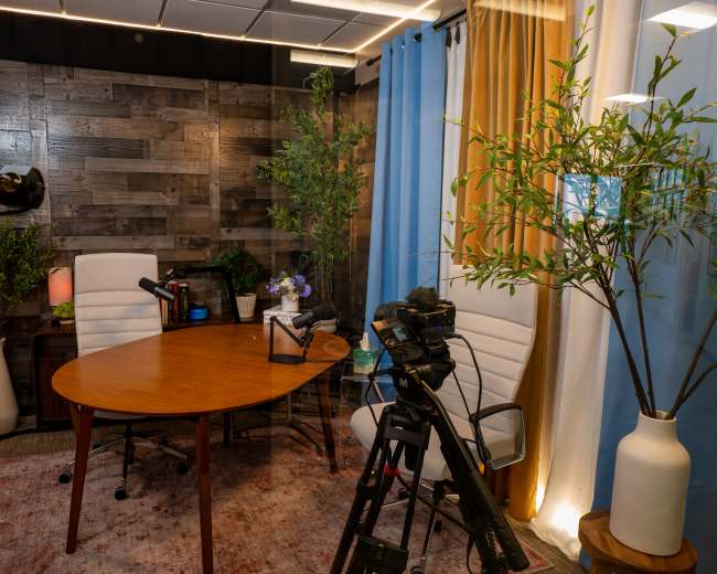 The image shows a small studio space with a wooden table surrounded by two white chairs, a camera on a tripod, and decorative plants, all under soft lighting.