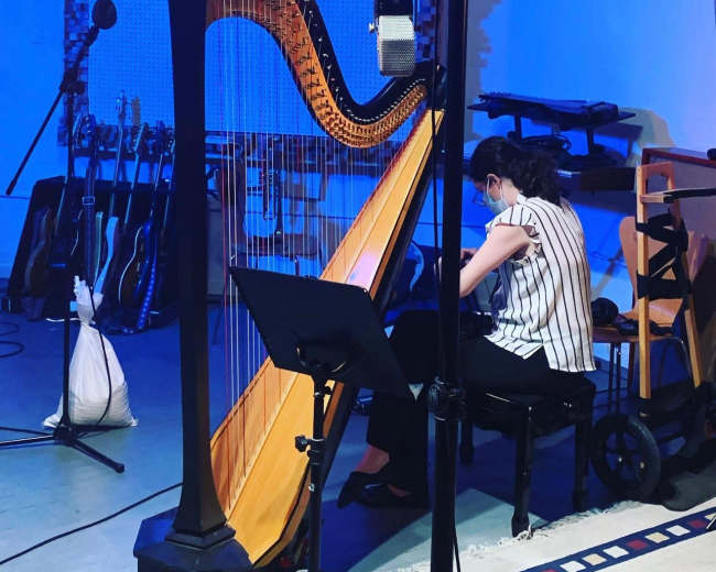 A musician plays a harp in a well-equipped studio surrounded by guitars and microphones.