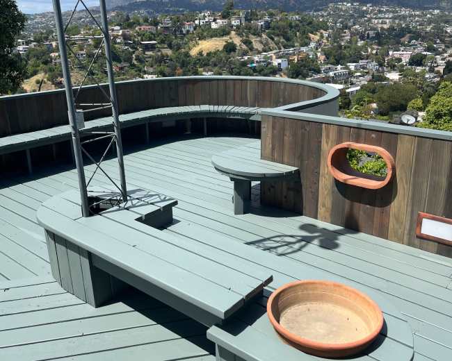 A wooden deck with a curved seating area, a fire pit, and a view of hills and buildings in the background.