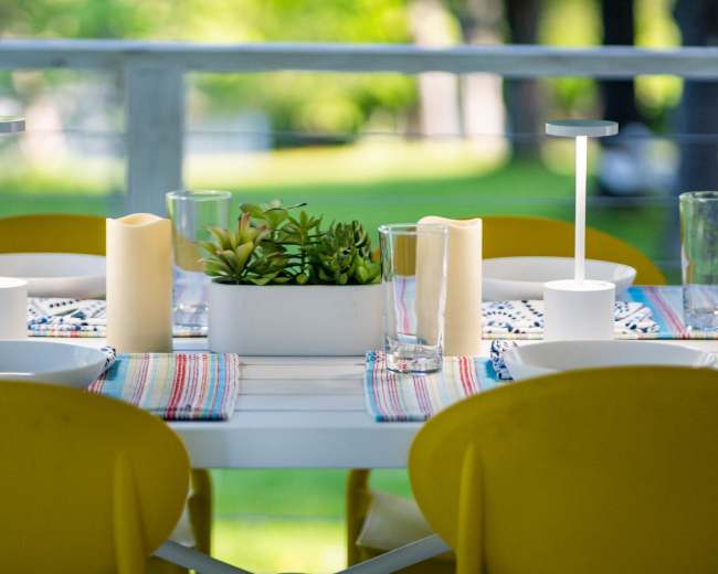 A dining table is set with yellow chairs, white dishes, glasses, and decorative candles, along with a potted plant centerpiece on a striped tablecloth.