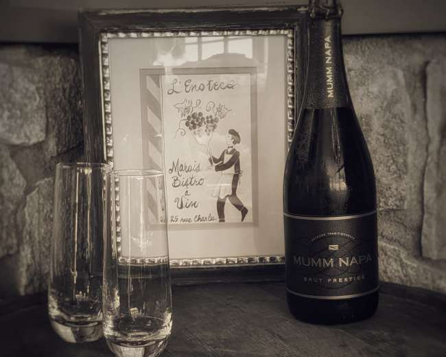 A bottle of Mumm Napa Brut Prestige is displayed on a wooden barrel alongside two fluted glasses and a framed art piece on the wall.