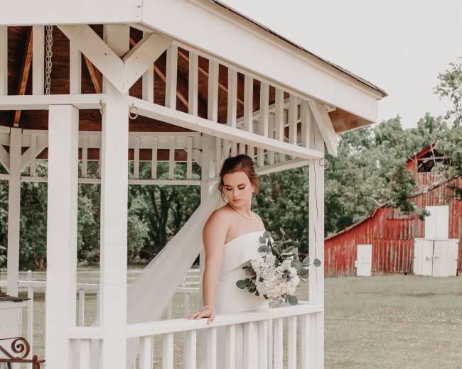 A bride in a white dress stands in a gazebo, holding a bouquet and looking down, with a red barn visible in the background.