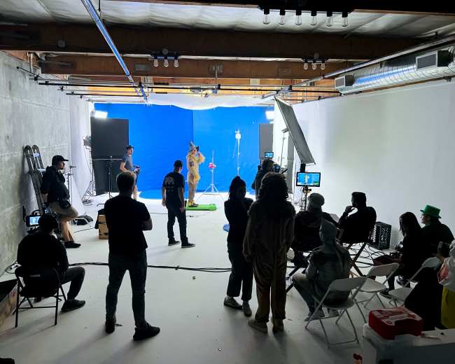 A group of people is filming a scene in a studio with a blue backdrop, while others observe from seated positions.