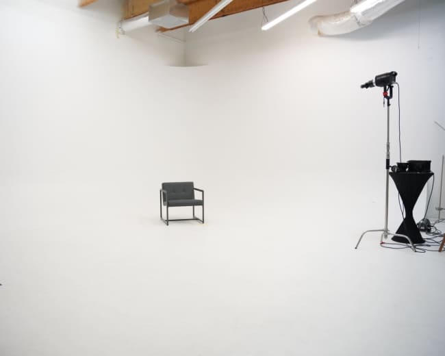 The image shows an empty, well-lit studio with a single gray chair in the center and photography equipment in the background.