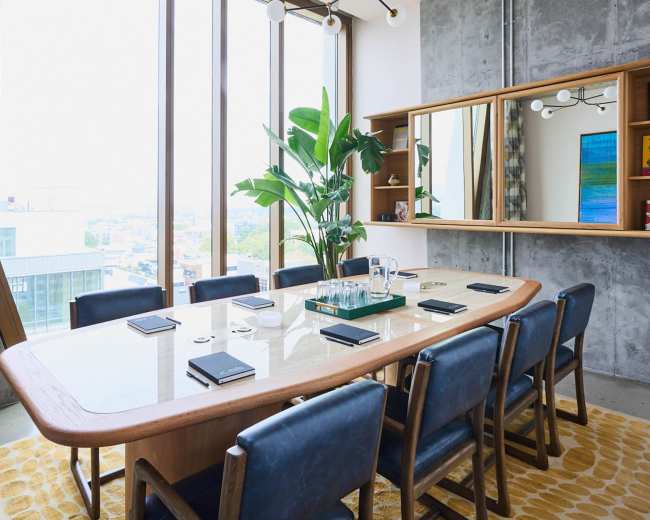 The image shows a modern conference room with a large wooden table surrounded by blue chairs, large windows offering a city view, and a potted plant in the corner.