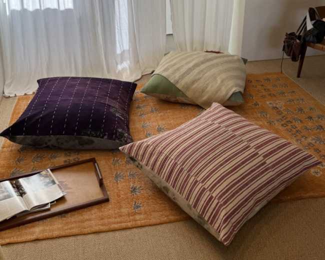 The image shows a cozy room with three large pillows arranged on a rug, alongside a wooden tray and an open magazine.