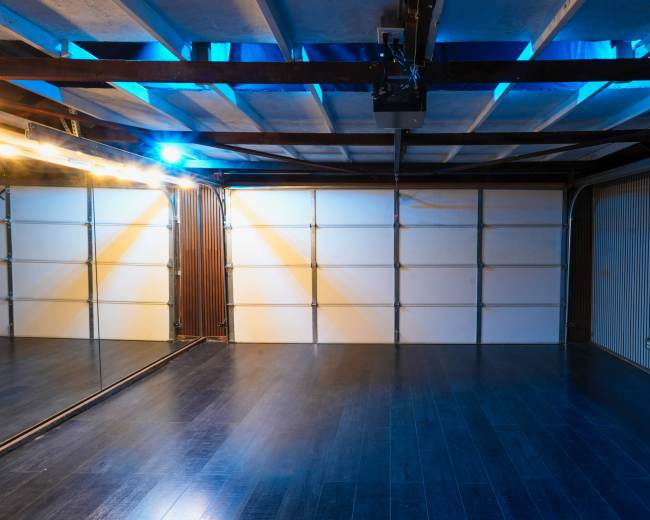 The image shows an empty studio space with mirrored walls, wooden flooring, and blue lighting.
