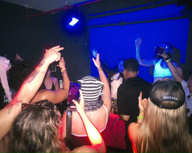 A group of people with their arms raised is gathered around a DJ at a lively indoor party, surrounded by colorful lights.