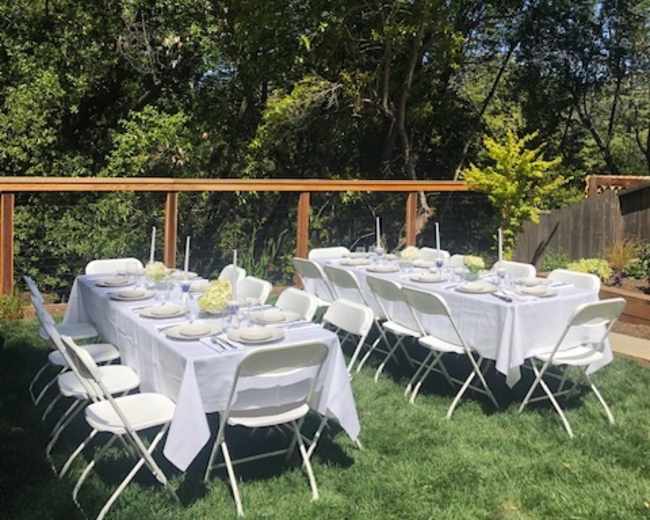 A long rectangular table is set with white tablecloths and dishes under a wooden deck surrounded by greenery.