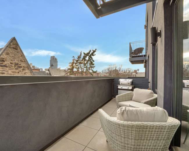 The image shows a modern balcony with two wicker chairs, overlooking rooftops and trees against a clear blue sky.