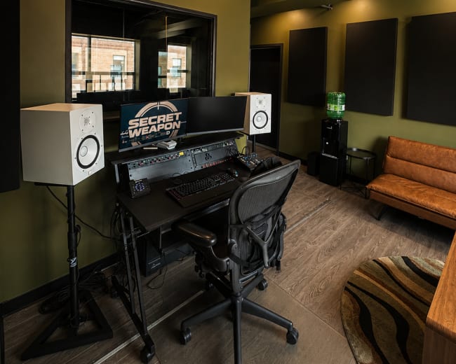 The image shows a music production studio featuring a desk with audio equipment, two speaker monitors on stands, and a comfortable seating area against a green wall.