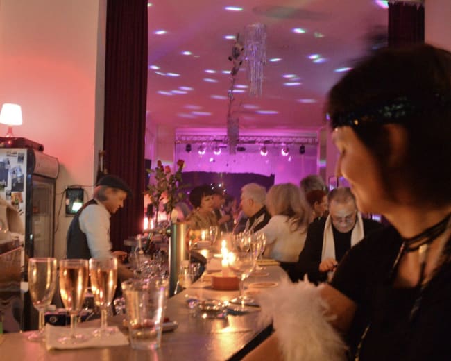 A lively gathering is depicted at a bar, with guests enjoying drinks and colorful lights illuminating the scene.