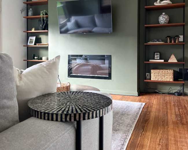 The image shows a modern living room featuring a mounted television, a linear fireplace, and open shelving with decorative items, all arranged around a sectional sofa.