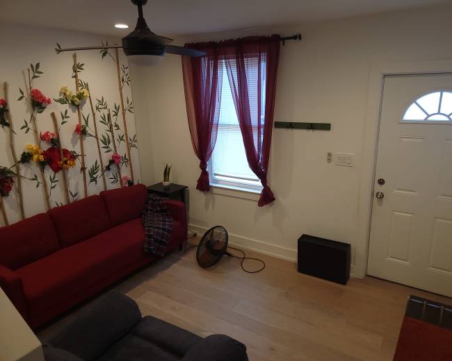 The image shows a living room with a red couch, a door with a window, a small black cabinet, and decorative wall panels featuring flowers.