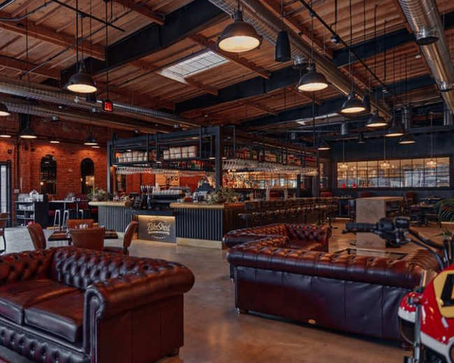 The image shows a modern bar interior featuring leather couches, a long bar with shelves of liquor, and an industrial-style design with exposed beams and large windows.