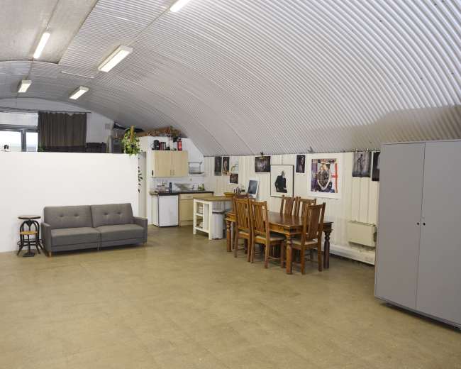The image shows a spacious, curved-ceiling room featuring a gray couch, a wooden dining table with chairs, a kitchenette area, and several framed pictures on the walls.