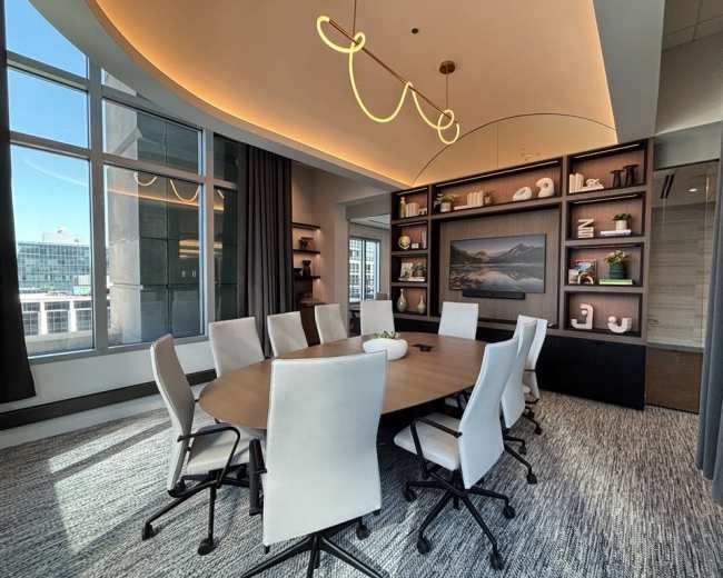 A modern conference room features a large round table surrounded by white chairs, with a gallery-style display and large windows letting in natural light.