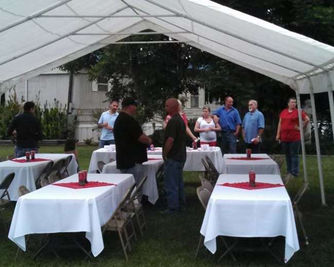 A group of people is gathered under a large white tent with tables covered in white cloths and red centerpieces, set up in a grassy area.