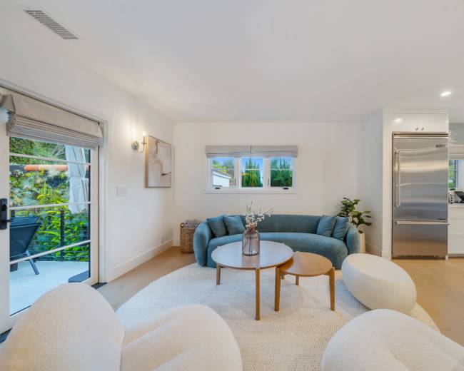 The image shows a modern living room with a blue sofa, two round tables, and white chairs, featuring large windows that overlook a garden.