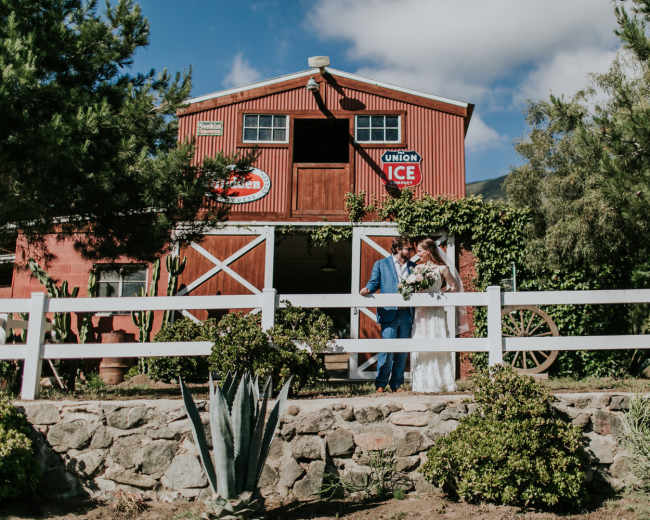 A couple stands together in front of a red barn adorned with signs, surrounded by greenery and decorative plants.