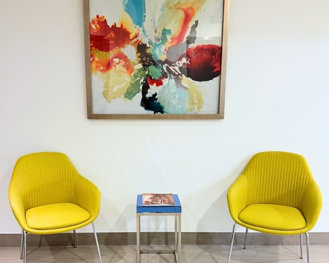 The image shows two yellow chairs positioned on either side of a small metallic table, with a colorful abstract painting hanging above them on a light-colored wall.