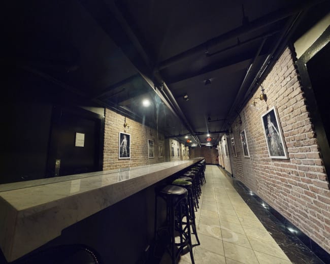 The image shows a long, narrow bar area with a marble counter and black stools, flanked by exposed brick walls and framed photographs.