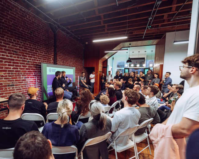 A large audience sits in a room with exposed brick walls, listening to speakers on a stage during an event.