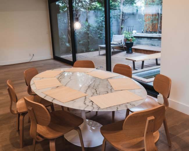 A round marble dining table with six wooden chairs is positioned in a room that features large glass doors opening to a patio view.