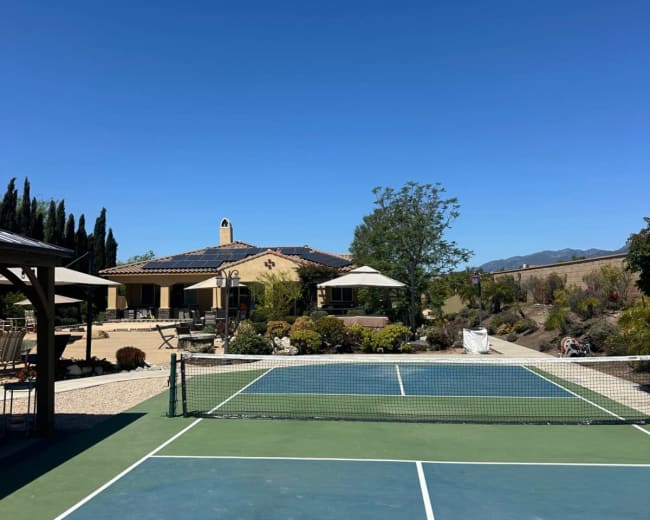 The image shows a tennis court in the foreground with a house and landscaped garden in the background under a clear blue sky.