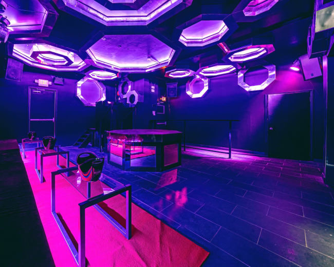 The image shows a dimly lit interior of a modern lounge with purple and blue lighting, featuring hexagonal light fixtures and a central table surrounded by empty seating.