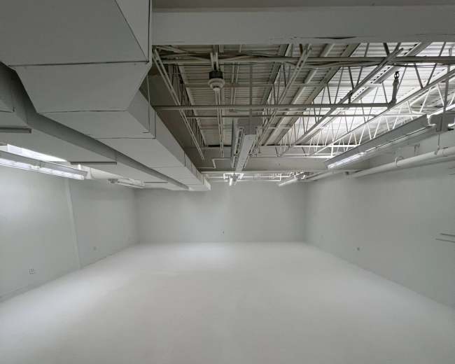 The image shows a large, empty room with white walls and a white floor, illuminated by overhead lights.