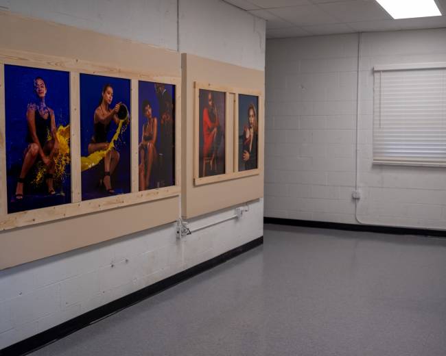 The image shows a hallway with four framed photographs of individuals displayed on one wall, while the opposite wall features a window with blinds.