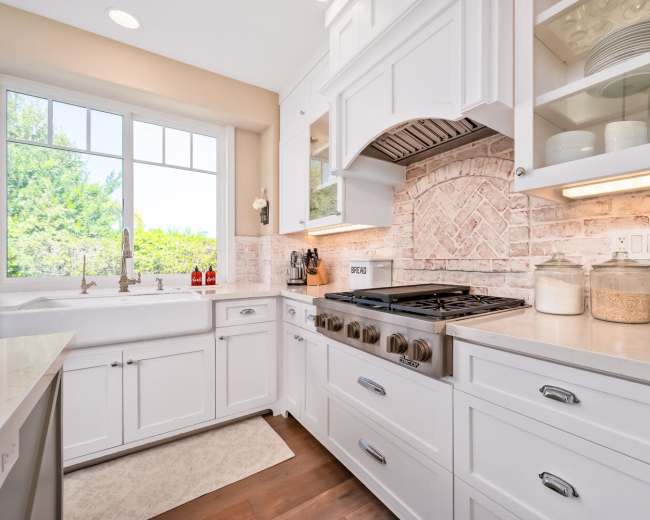 A well-organized kitchen with white cabinetry, a farmhouse sink, and a gas range under a decorative hood, along with jars of ingredients displayed on the countertop.