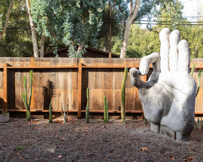 A large concrete hand sculpture stands in a gravel yard surrounded by various cacti against a wooden fence.