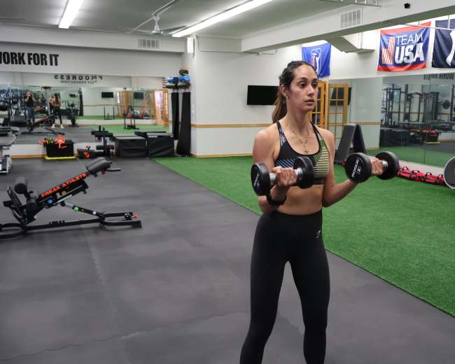 A woman lifts dumbbells in a fitness gym equipped with various exercise equipment.