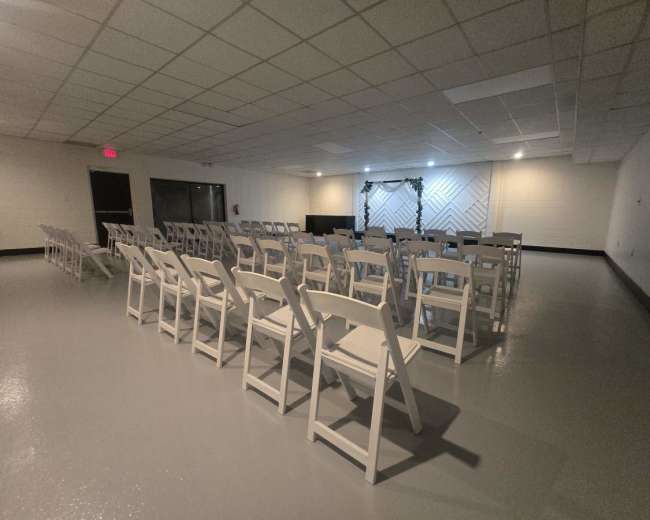The image shows a spacious, empty room set up with rows of white folding chairs facing a decorative backdrop.