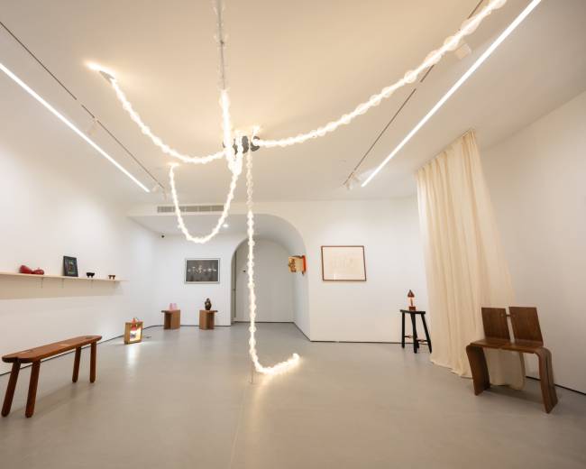 The image shows an art gallery with minimalist decor, featuring a series of light strands hanging from the ceiling and various sculptures displayed on walls and pedestals.