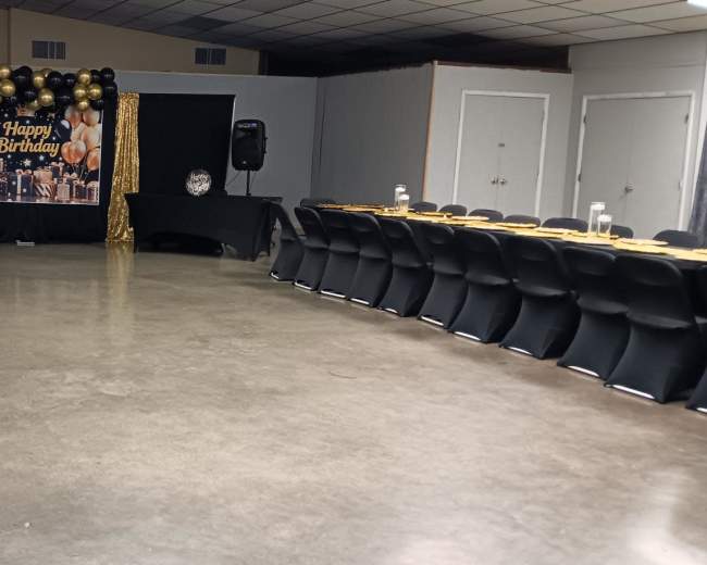 The image shows a large indoor event space set up for a birthday celebration, featuring rows of tables with black tablecloths, gold accents, and a decorated backdrop with balloons and a "Happy Birthday" sign.