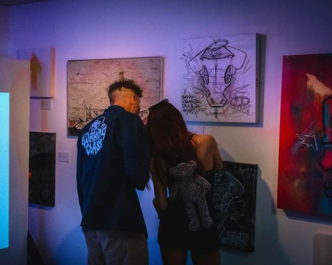 Two people examine artwork on the wall in a gallery, while a projection of a weapon is displayed on a nearby surface.