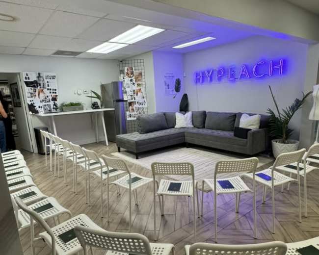The image shows a modern room set up with a gray sectional sofa, a neon sign that reads "HYPERACH," and white chairs arranged in rows, suitable for a meeting or event.
