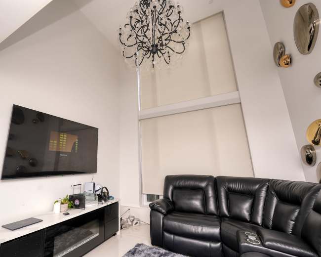 The image shows a modern living room featuring a black leather sofa, a large flat-screen TV mounted on a white wall, and a decorative chandelier hanging from the ceiling.